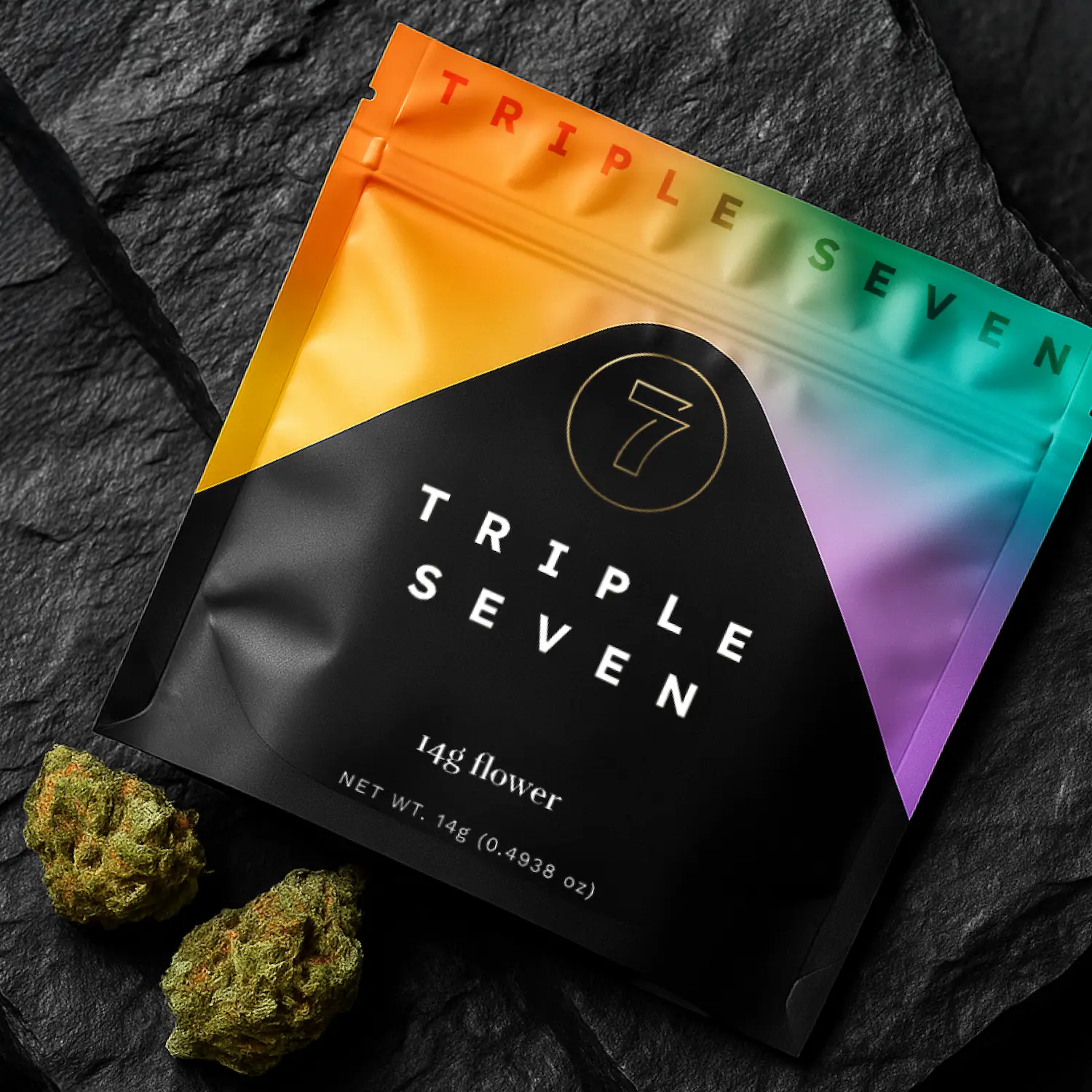 Triple Seven cannabis flower packaging in a sleek black pouch with a vibrant rainbow gradient top, displayed on a dark stone surface beside two buds of cannabis.