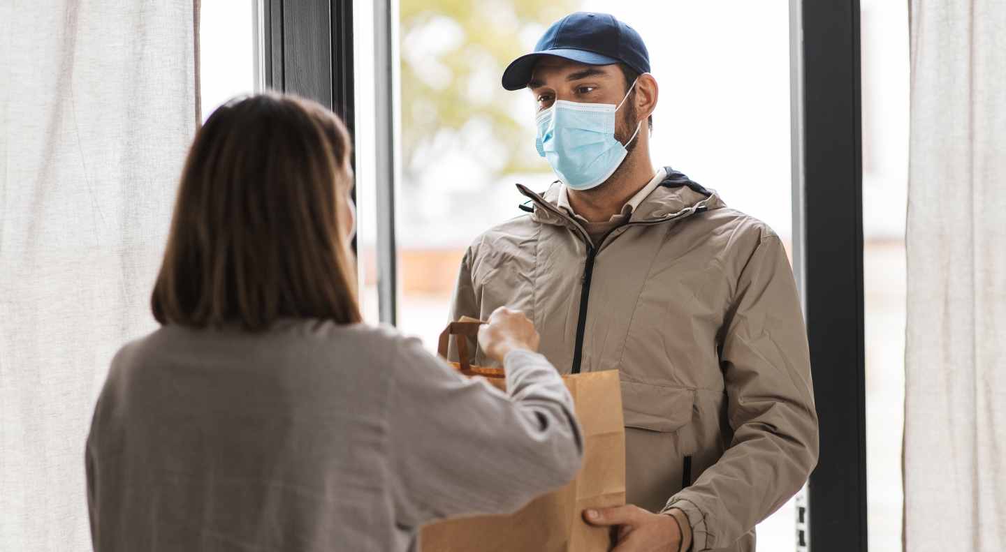 A delivery person wearing a beige jacket, blue cap, and face mask hands a brown paper bag to a woman at her door. The woman, seen from behind, reaches out to receive the package. The setting includes a bright indoor space with large windows and curtains.