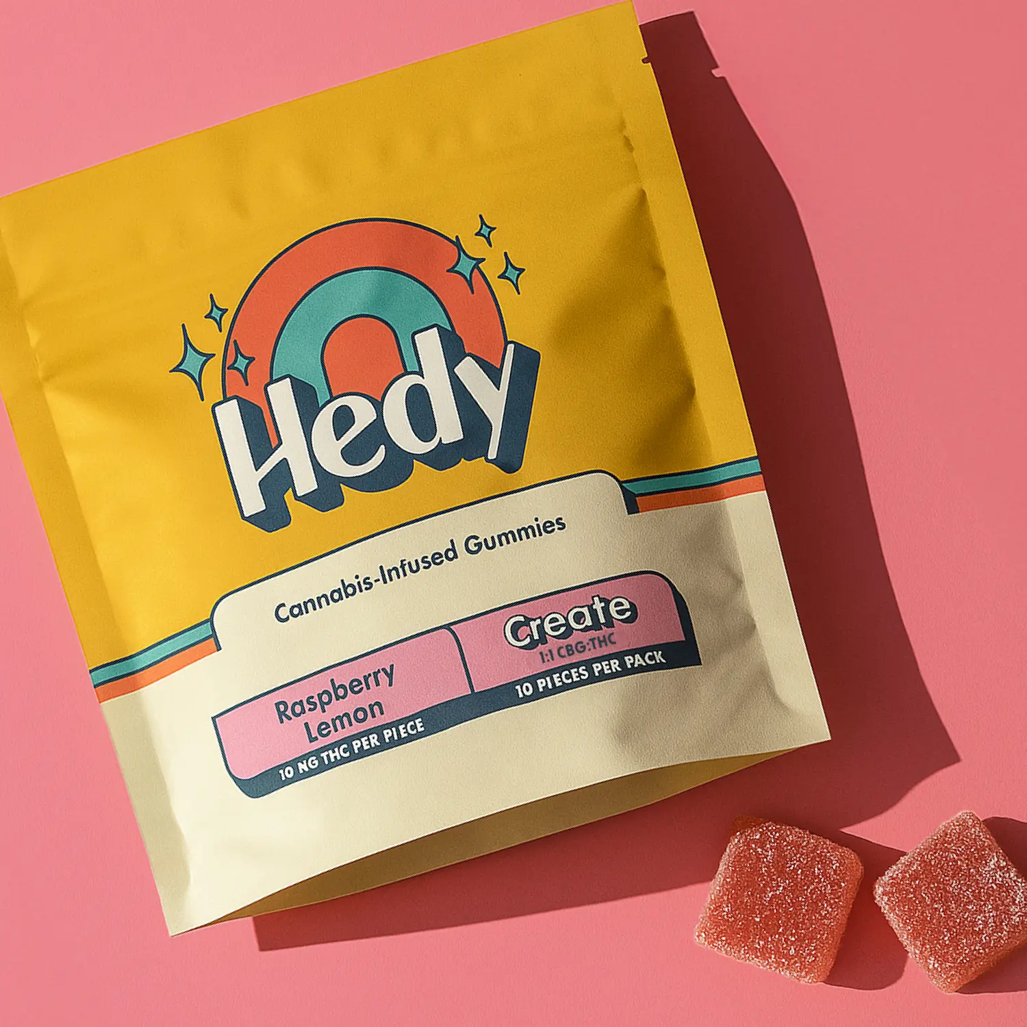 Hedy Raspberry Lemon cannabis-infused gummies in bright retro packaging with rainbow design, photographed on a pink backdrop with two sugar-dusted gummies beside the bag.